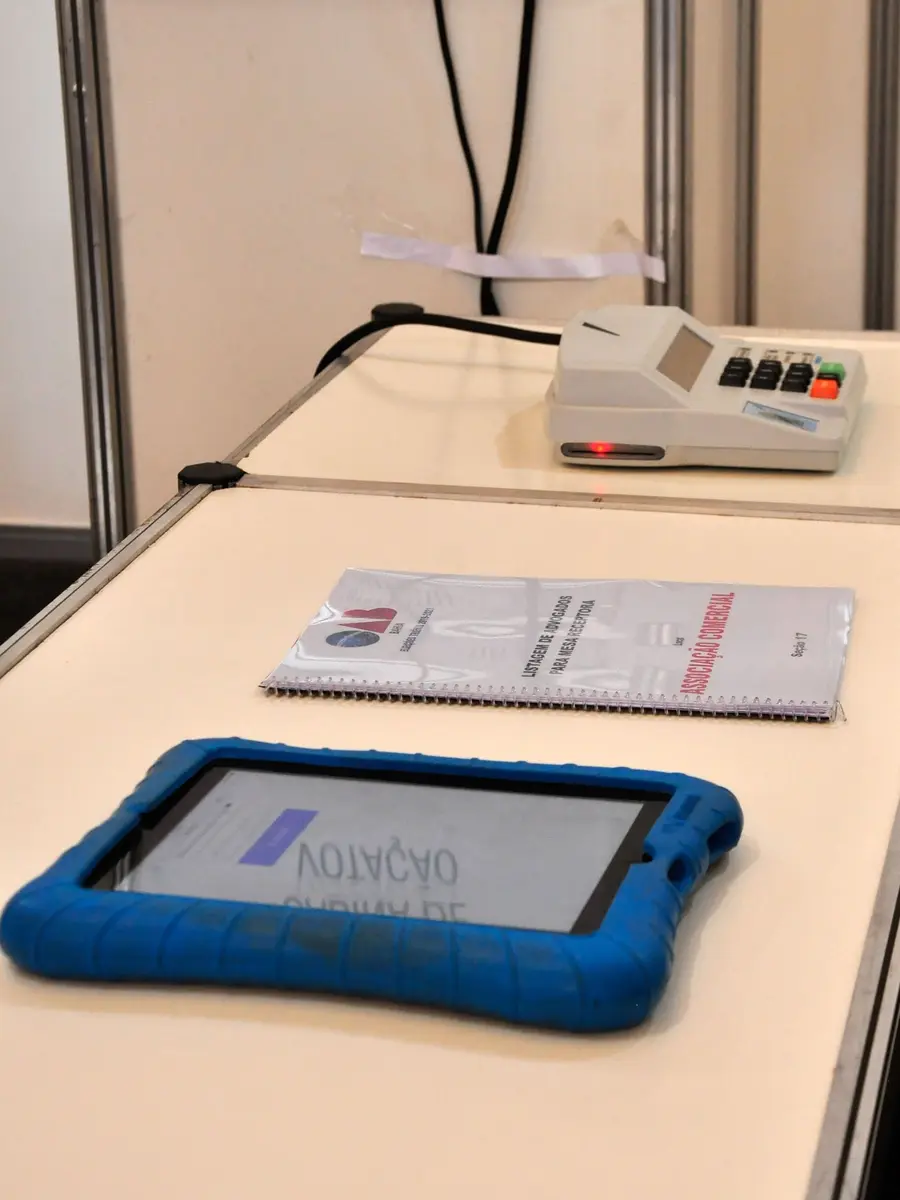 Tablet de capa azul sobre a mesa de votação, ao lado do terminal do mesário e da listagem de advogados da seção